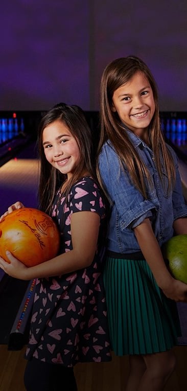 Two girls standing back to back holding bowling balls and smiling