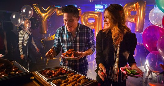 Two party guests grabbing food at the buffet with balloons in the background