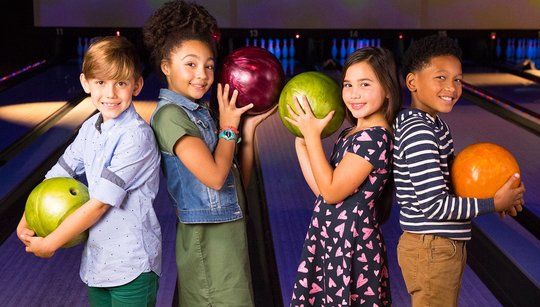 Four kids standing in front of bowling lanes, holding bowling balls, and smiling