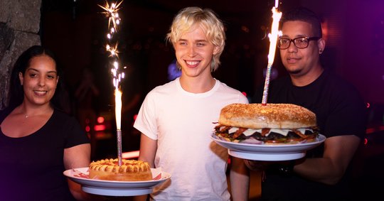 Boy in between two Bowlero servers, holding giant hamburger and pizza item with sparks coming out of them