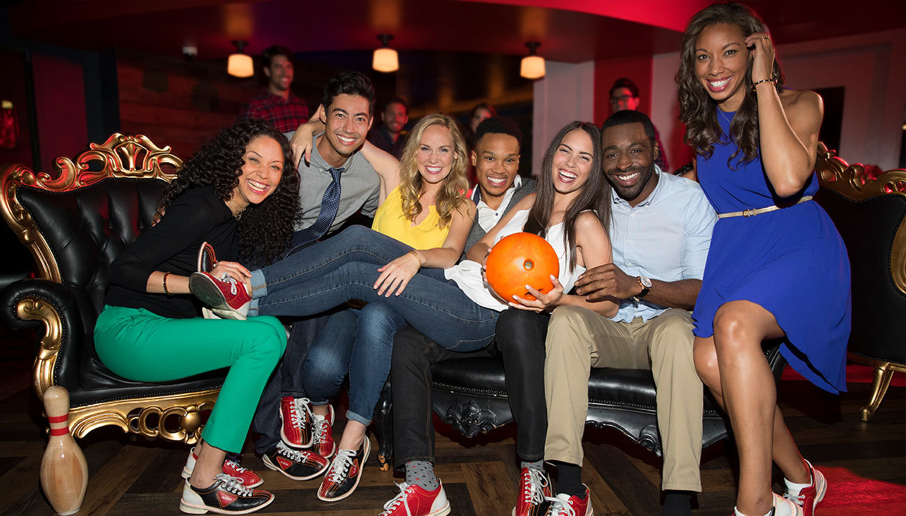 Seven smiling people sitting on a couch and one is holding a bowling ball