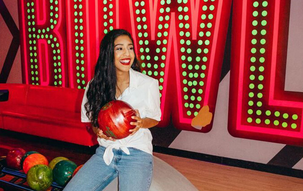Young woman holding a bowling ball and posing in front of a Bowl Sign